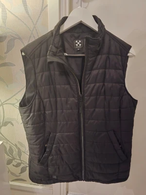 Svart dunväst  - Snygg svart dunväst från JH Collection med quiltat mönster och hög krage. Västen har dragkedja framtill och två fickor med dragkedja. Perfekt att slänga över en hoodie eller tröja för extra värme.