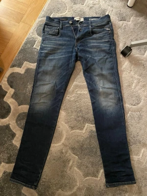 Replay Anbass W28 L32 - Snygga mörkblå Replay Anbass jeans med klassisk femficksdesign och diskreta slitningar framtill. Jeansen har smal passform och raka ben, tillverkade i mjukt jeansmaterial med stretch för extra comfort