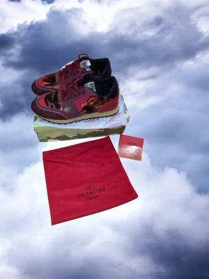 Valentino Rockrunner röda sneakers - Säljer ett par Valentino Rockrunner sneakers i rött och svart med camouflage-mönster. Skorna har snörning, rund tå och detaljer i mocka och skinn. Snygg design med beige sula och svarta detaljer. Kommer med originalpåse och box.