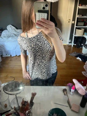 Leopardmönstrad topp med offshoulder - Säljer en snygg leopardmönstrad topp med offshoulder-design. T-shirten har korta ärmar och en lös passform, perfekt för dig som gillar trendiga och bekväma plagg. Materialet känns mjukt och lätt mot huden.