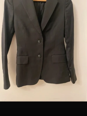 Svart klassisk kavaj med knappar - Stilren svart kavaj med två knappar framtill och klassiska slag. Kavajen har två fickor med lock och är helfodrad. Perfekt för en clean och tidlös look. Passar till allt från skjorta till t-shirt.