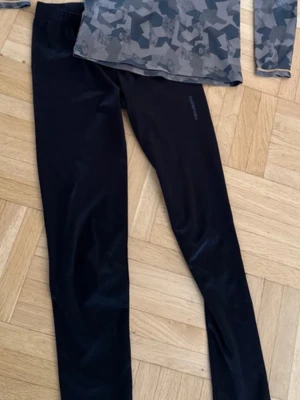 Underställ från Tenson, camo & svart 158/164 - Snyggt träningsset från Tenson med långärmad tröja i grått camomönster och svarta tights. Tröjan har guldfärgade sömmar och rund hals. Materialet är stretchigt och andas, perfekt för löpning eller gym. Tightsen är helsvarta med diskret logga på benet.
