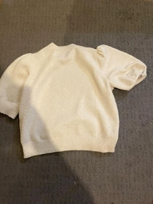 Krämvit puffärm tröja från H&M - Säljer en krämvit tröja från H&M med korta puffärmar och ribbade muddar. Tröjan har en mjuk, stickad struktur och en avslappnad passform som ger en trendig look. Perfekt att styla med jeans eller kjol för en cool outfit.