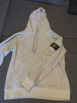 Grå hoodie från Stone Island - Snygg grå hoodie från Stone Island med klassisk logotyp-patch på vänster ärm. Hoodien har dragsko i huvan och ribbade muddar vid ärmslut och nederkant. Perfekt för en avslappnad och trendig stil. Pris kan diskuteras vid snabb affär 