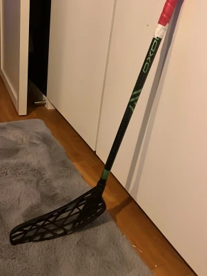 MINI OXDOG innebandyklubba svart/grön - Säljer en MINI OXDOG innebandyklubba i svart med gröna detaljer. Klubban har ett svart blad med öppet mönster och skaftet är lindat med rött och vitt grepp. Perfekt för dig som gillar innebandy och vill ha en lätt och smidig klubba.