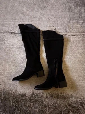 Svarta höga mockastövlar med klack - Snygga svarta stövlar i mocka med rund tå och låg klack. Stövlarna har dragkedja på insidan och en stilren design som passar till många outfits. Perfekta för höst och vinter när du vill ha både stil och komfort.