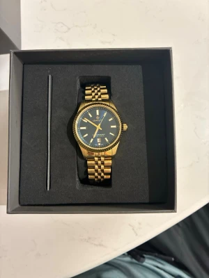 Guld Unisex klocka från GANT - Den här guldiga GANT-klockan är en stilren favorit som passar alla. Med ett ur på 37,5 mm har den en perfekt balanserad storlek som känns lika rätt till vardags som vid finare tillfällen. Den navyfärgade urtavlan ger ett lugnt och elegant intryck som matchar alla stilar. En klocka som är enkel att bära, lätt att tycka om och svår att tröttna på. Aldrig använd.  Klockan levereras i orginalförpackning med extra länkar om man vill vidga den samt kvitto på klockan mm.