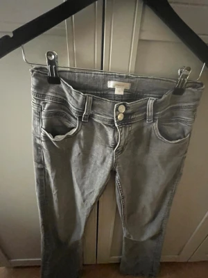 Grå bootcut jeans med dubbla knappar - Snygga grå jeans med bootcut-modell och dubbla knappar i midjan. Jeansen har klassiska fickor fram och bak samt markerade sömmar. Perfekta för dig som gillar en avslappnad men trendig look. Med en silver stjärna på ena fickan 