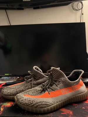 Adidas Yeezy Boost 350 V2 med orange stripe - Säljer ett par Adidas Yeezy Boost 350 V2 sneakers med gråmelerad ovandel i stickad textil och en tydlig orange stripe på sidan. Skorna har snörning och en genomskinlig, ribbad sula som ger en futuristisk vibe. Perfekta för dig som gillar streetwear och vill sticka ut.