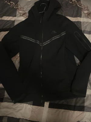 Svart Nike hoodie med dragkedja - Säljer en svart hoodie från Nike med dragkedja framtill och huva. Tröjan har en diskret Nike-logga på bröstet och en snygg diagonal detalj i grått över bröstet. Ärmarna är långa och det finns en ficka med dragkedja på ena ärmen. Perfekt för en sportig och avslappnad stil.