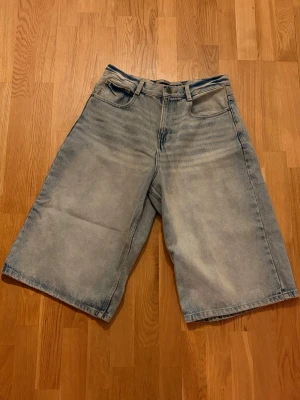Ljusblåa loose jeansshorts - Säljer ett par ljusblåa jeansshorts med loose fit och fem klassiska fickor. Shortsen har bälteshällor och en avslappnad, rak passform som går ner till knäna. 