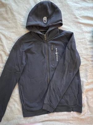 Mörkblå Sail Racing hoodie - Mörkblå hoodie från Sail Racing med dragkedja framtill och logga på bröstet. Huva med tryckt detalj och två sidofickor med dragkedja. Tillverkad i mjukt material som känns skönt mot huden. Perfekt för en avslappnad och sportig stil.