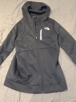 Mörkgrå ziphoodie från The North Face - Snygg mörkgrå ziphoodie från The North Face med huva och dragkedja framtill. Tröjan har en bröstficka med dragkedja och logga på både fram- och baksidan. Tillverkad i mjukt syntetmaterial och har en clean, sportig look. En allmänt skön vardaglig tröja som passar perfekt i skolan eller ute med vänner. Tröjan är aldrig använd och jag säljer den pga att jag fick den i presen och den var tyvärr för liten. Storleken är XXL i barnstorlek men jag skulle beskriva den som en XS/S i vuxenstorlek.