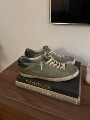 Gröna Golden Goose sneakers - Säljer ett par snygga gröna sneakers från Golden Goose med vita snören och svart hälparti. Skorna har klassisk låg profil, rund tå och är tillverkade i mocka med detaljer i skinn. Perfekta för dig som gillar en avslappnad och trendig stil.