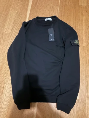 Svart sweatshirt från Stone Island - Svart sweatshirt från Stone Island med klassisk rund hals och ribbade muddar. Tröjan har den ikoniska Stone Island-loggan på vänster ärm. Tillverkad i mjuk bomull för skön känsla och stilren look. Perfekt för dig som gillar streetwear och enkel design. Helt ny aldrig använt. Stl XL