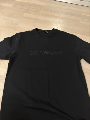 Svart Emporio Armani t-shirt - Snygg svart t-shirt från Emporio Armani med diskret logga tryckt på bröstet. Klassisk rund halsringning och korta ärmar. Perfekt för dig som gillar stilrena och exklusiva plagg med en modern touch.