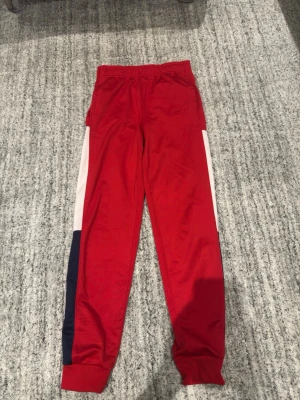 Röda mjukisbyxor med färgblock - Snygga röda mjukisbyxor med elastisk midja och ribbade benslut. Byxorna har vita och marinblå sidopaneler som ger en sportig vibe. Perfekta för chill eller träning. Tillverkade i mjukt syntetmaterial för maximal komfort.