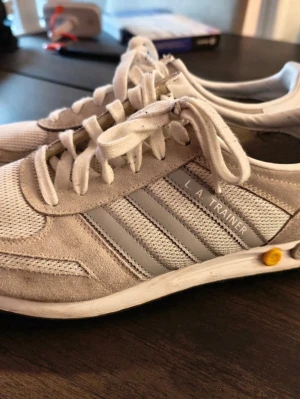 Adidas L.A. Trainer sneakers grå/vit - Snygga Adidas L.A. Trainer sneakers i grått och vitt med klassiska tre ränder på sidan. Ovandelen är i mesh och mocka för en sportig look. Skorna har snörning och platt sula med detaljer i gult vid hälen. Perfekta för en avslappnad stil.