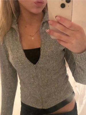 Grå ribbad ziptröja med hög krage - Säljer en grå ribbad ziptröja med hög krage och långa ärmar. Tröjan har en figurnära passform och dragkedja framtill. Perfekt att slänga på sig över ett linne eller t-shirt för en chill look.