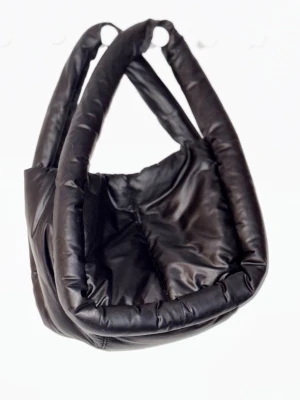 Svart puffad handväska från DAY ET - Trendig svart puffad handväska från DAY ET med breda, vadderade handtag och mjuk form. Väskan har innerficka med dragkedja och är tillverkad i syntetmaterial. Perfekt för dig som vill ha en cool och modern accessoar till din outfit. 