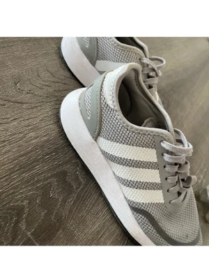 Gråa Adidas sneakers med vita ränder - Snygga gråa Adidas sneakers med klassiska vita ränder på sidorna. Ovandelen är i meshmaterial för extra andningsförmåga och skorna har snörning framtill. Perfekta för en sportig och stilren look. Fint skick.