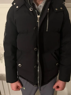 Svart pufferjacka från Moose Knuckles - Säljer en svart pufferjacka från Moose Knuckles med stora silvriga dragkedjor, knappar och ikonisk logga på ärmen. Jackan har huva, flera fickor och en schysst oversized look. Perfekt för kalla dagar och riktigt snygg till streetwear. ( pälsen finns att få med )