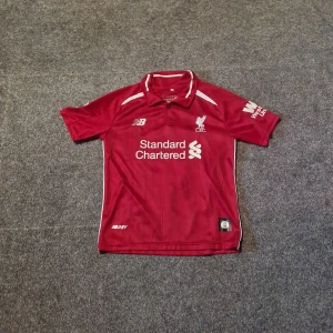 Liverpool M.Salah matchtröja & shorts - Liverpool FC matchtröja med M.Salah och nummer 11 på ryggen, tillverkad i röd polyester med vita detaljer och klubbmärke på bröstet. Säljer även tillhörande röda shorts med klubbmärke och New Balance-logga. Perfekt för dig som vill köra full kit på träningen eller rocka på skolan.