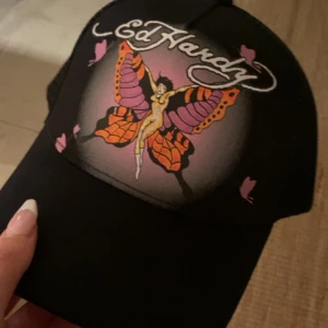 Svart Ed Hardy keps med fjärilstryck - Säljer en svart keps från Ed Hardy med ett färgstarkt tryck av en fjäril och en kvinna på framsidan. Kepsen har coola lila och orange detaljer och ett stort Ed Hardy-logo ovanför motivet. Perfekt för dig som gillar statement-plagg och unika prints. 