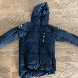 Svart pufferjacka Sail Racing - Säljer en svart pufferjacka från Sail Racing med huva och dragkedja framtill. Jackan har logga på bröstet och reflekterande detaljer på ärmarna. Perfekt för kalla dagar med sin vadderade design och långa ärmar. Snygg och funktionell för vintern.