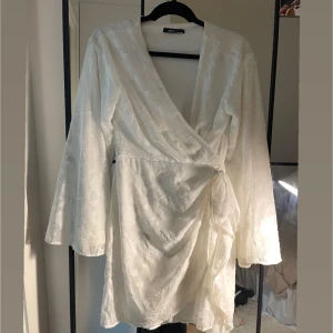 Vit omlottklänning från Gina Tricot - Superfin vit omlottklänning från Gina Tricot med subtilt blommönster i tyget. Klänningen har långa ärmar, v-ringning och knyt i midjan för en snygg passform. Perfekt för dig som gillar en fräsch och stilren look.