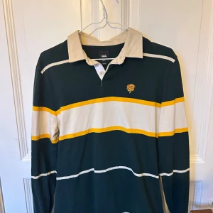 Randig pikétröja från Vans - Snygg långärmad pikétröja från Vans i mörkgrönt med breda vita och gula ränder. Klassisk krage i beige och broderad ros på bröstet. Perfekt för dig som gillar retrostil och streetwear.