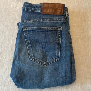 Blå jeans från Tiger Of Sweden - Snygga blå jeans från Tiger of Sweden, modell LEA, tillverkade i Italien. Klassisk femficksdesign med bruna sömmar och läderpatch bak i midjan. Jeansen har en rak passform och är gjorda i slitstarkt denimtyg. Perfekta till sneakers eller boots. Storlek 29/32. Inget FAST pris!