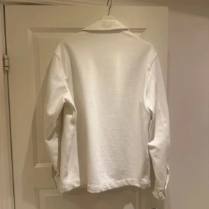 Vit overshirt från Zara - Snygg vit overshirt från Zara med klassisk krage och stora bröstfickor. Jackan har knäppning framtill med vita knappar och långa ärmar med manschett. Perfekt att slänga över en hoodie eller t-shirt för en clean look.