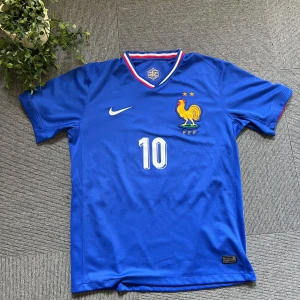 Blå Frankrike Mbappé fotbollströja Nike - Säljer en officiell blå Frankrike fotbollströja från Nike med Mbappé och nummer 10 på ryggen. Tröjan har korta ärmar, broderat landslagsmärke med tupp och två stjärnor, samt vita och röda detaljer vid kragen. Tillverkad i lätt polyester för maximal komfort på planen. Storlek L men sitter som M