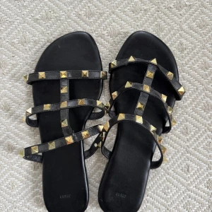 Svarta sandaler med guldfärgade nitar - Snygga svarta sandaler med flera remmar dekorerade med guldfärgade pyramidnitar. Platt sula och öppen tå ger en chill vibe. Remmarna är i skinnimitation och sulan har ett geometriskt mönster undertill. Ser ut som klackar men är flats. Har endast testat de på en gång så skulle säga helt nytt skick, säljer pga inte kommit till användning. Skriv för mer info 🩷