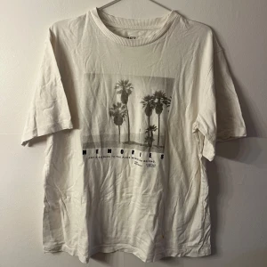 Vit t-shirt med palmtryck - Vit t-shirt med coolt svartvitt palmtryck och texten 'MEMORIES' på framsidan. Klassisk rund hals och korta ärmar. Perfekt för dig som gillar chill vibes och grafiska prints. Mjuk bomullskänsla och relaxed fit.