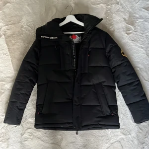 Svart pufferjacka från Zavetti Canada - Säljer en svart pufferjacka från Zavetti Canada med huva och feta broderade loggor på bröstet och ärmen. Jackan har dragkedja, stora fickor och quiltad design. Perfekt för kalla dagar när du vill hålla stilen och värmen.