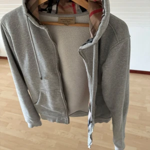 Grå hoodie från Burberry med dragkedja - Snygg grå hoodie från Burberry med klassiskt rutigt foder i huvan. Tröjan har dragkedja framtill, snörning i huvan och två fickor. Perfekt för dig som gillar stilren design med en touch av lyx. Materialet är mjuk bomull och passformen är normal.