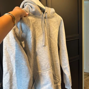 Grå hoodie med huva och ficka - Säljer en enkel och chill grå hoodie med stor ficka framtill och snörning vid huvan. Perfekt för att ta på sig när det är lite kyligt ute. Mjuk och skön bomullskänsla, långärmad och loose fit. Passar dig som gillar basic och comfy stil.