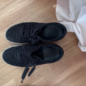 Svarta sneakers i mocka från Brixtad - Svarta sneakers från Brixtad i mjuk mocka med klassisk låg profil och snörning. Skorna har en beige sula som ger en snygg kontrast mot det svarta ovandelen. Perfekta för dig som gillar en stilren och enkel look. Ena snöret är lite långt men inget som påverkar 