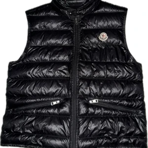 Svart dunväst från Moncler - Svart dunväst från Moncler med glansig finish och horisontella quiltade paneler. Västen har två snedställda fickor med dragkedja och klassisk Moncler-logga på bröstet. Hög krage och hel dragkedja framtill. Perfekt för lager-på-lager under kyliga dagar. Storlek 2 motsvarar small äkta såklart.