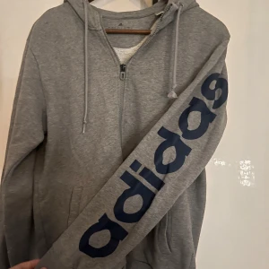 Grå hoodie från Adidas med tryck - Säljer en grå hoodie från Adidas med dragkedja och huva. Stort mörkblått Adidas-tryck längs ena ärmen och liten logga på huvan. Tröjan har ribbade muddar och fickor framtill. Lite stor i storlek?