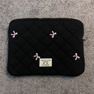 Datorfodral  - Quiltad svart datorväska från Kazum med ljusrosa broderade rosetter och dragkedja upptill. Väskan har en mjuk, vadderad känsla och ett litet tygmärke med texten 'make your day happier by kazum' på framsidan. Perfekt för att bära din laptop med stil.