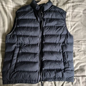 Mörkblå dunväst från Zara - Snygg mörkblå dunväst från Zara med quiltad design och hög krage. Västen har dragkedja framtill och två sidofickor. Perfekt att slänga över en hoodie för extra värme. Passar dig som gillar stilren och enkel look.