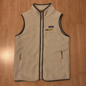 Beige fleeceväst från Patagonia M - Säljer en beige fleeceväst från Patagonia i storlek M. Västen har hög krage, tre fickor med dragkedja och snygga kontrastdetaljer i mörkbrunt. Perfekt för lager-på-lager och outdoor-vibe. Klassisk Patagonia-logga på bröstet. Priset är inte hugget i sten!