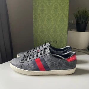 Gucci Ace  - Säljer de fetaste Gucci ace skorna på marknaden (Snygga Gucci sneakers i mörkgrå design med klassiska röda och blå ränder på sidan. Baksidan har metallicröd och metallicblå häl med Gucci-logga. Skorna har snörning, rund tå och vit platt sula. Materialet är präglat skinn med diskret GG-mönster.) UK10, passar mig som bär 43/44. Jättebra Skick, finns endast lite små slitningar (finns i bilderna) som inte syns vid användning. Box medföljer vid köp! Hör av dig vid köp/frågor (KÖP INTE DIREKT)