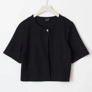 Svart croppad väst med knapp - Stilren svart croppad väst från Gina tricot med korta ärmar och rund hals. Västen har en enkel design med en guldfärgad knapp framtill. Perfekt för att lyfta din outfit med en minimalistisk touch.