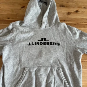 Grå hoodie från J.Lindeberg - Snygg grå hoodie från J.Lindeberg med svart broderad logga på bröstet. Klassisk känguruficka framtill och huva med dragsko. Perfekt för en chill och stilren look. Mjuk insida och normal passform. Snören kommer till. Ingen fläck eller hål