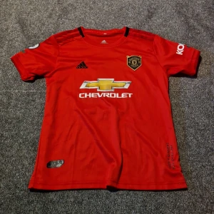 Manchester United Pogba matchtröja Adidas - Röd Manchester United fotbollströja från Adidas med Pogba och nummer 6 på ryggen. Tröjan har korta ärmar, svarta detaljer och klubbmärke på bröstet. Materialet är lätt och andas, perfekt för fotboll. Säljer även tillhörande svarta shorts med röda ränder och klubbmärke. 