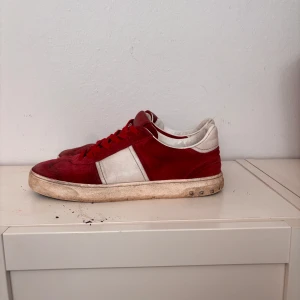 Valentino röda och vita sneakers mocka - Snygga sneakers från Valentino i röd mocka med vita detaljer och röda skosnören. Klassisk låg modell med rund tå och platt sula. Sidan har en vit panel och sulan är dekorerad med små nitar tyvärr har en par lossnat som du ser på bilden storlek 41-42 inget og kommer med